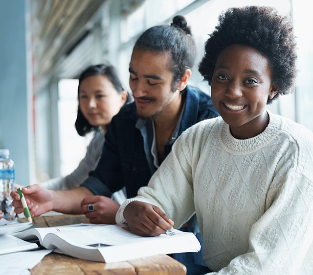 A young woman with a book is smiling. Two young people are sitting in the background.